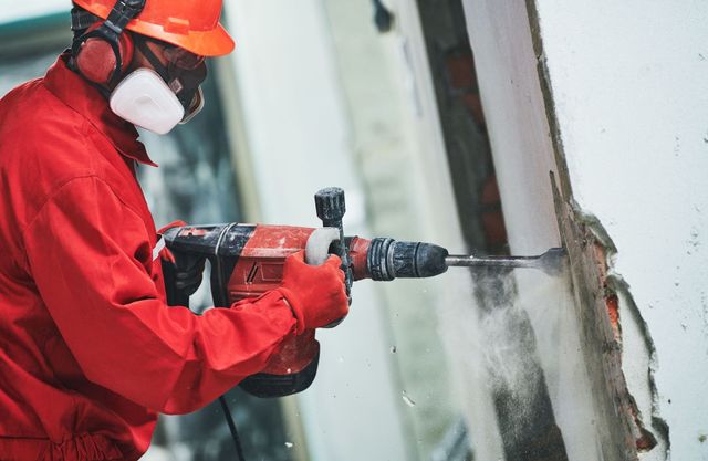 Worker in a hard hat and respirator using a rotary hammer to chisel out a wall