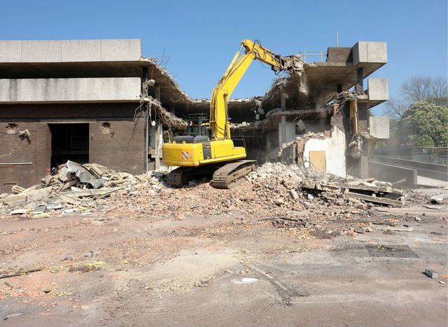 Excavator tearing down a concrete commercial building, with debris spread across the lot