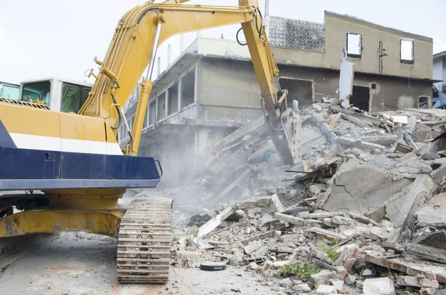 Excavator breaking apart a concrete building as dust rises from the rubble pile