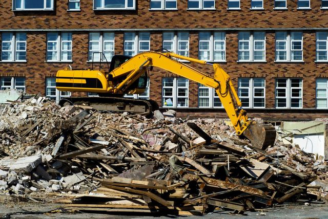 Yellow excavator sorting a pile of wood debris in front of a brick apartment building