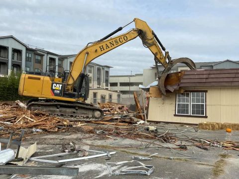 Yellow excavator demolishing a tan building, debris scattered. Buildings in background, overcast sky.