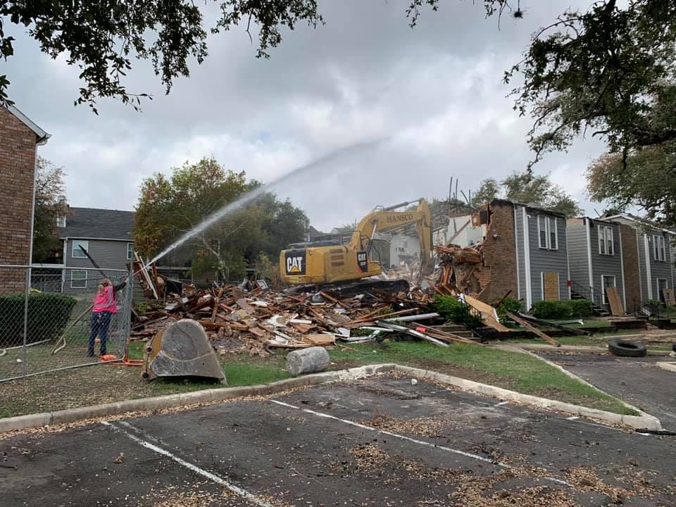 Worker spraying water for dust control while a Hansco excavator demolishes an apartment building