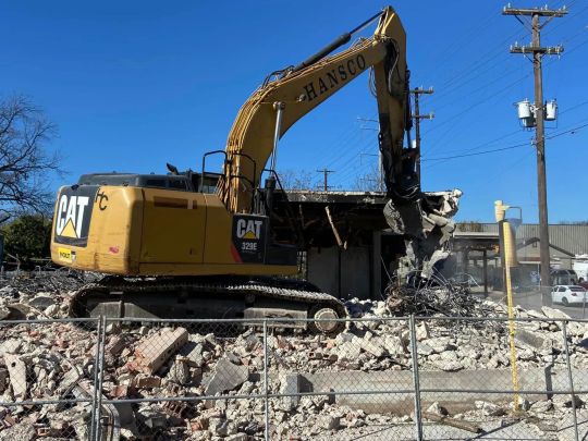 Yellow excavator demolishing a building on a sunny day. Bricks and debris on the ground.