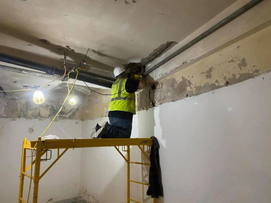 Worker on scaffolding drills into ceiling, wearing safety gear in a room with exposed pipes and damaged walls.