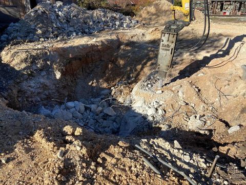 A backhoe breaking up concrete in an excavation site. Light brown dirt and debris surround the hole.