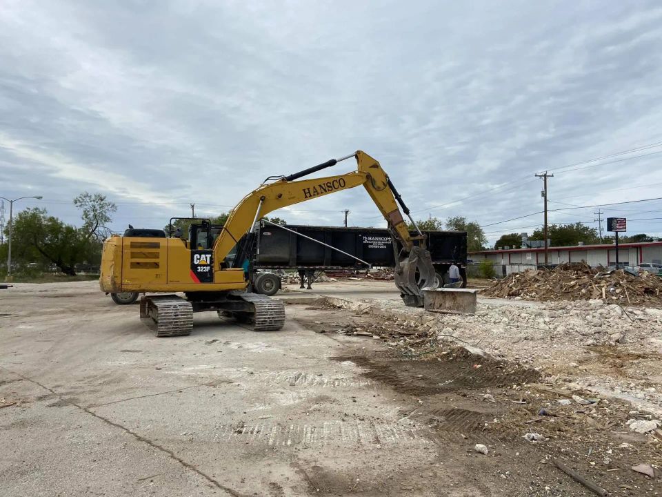 Excavator loading demolition debris into a black trailer on a cloudy day