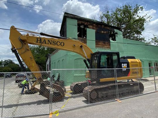 Yellow excavator demolishing a green building, fenced off, with a worker nearby.