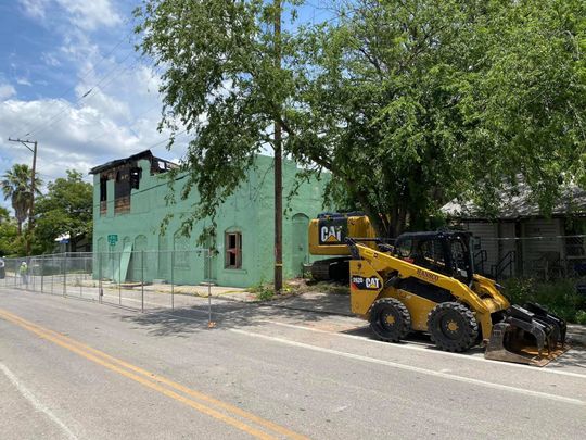 Yellow CAT skid steer on the street beside a fire-damaged green building behind a chain-link fence.