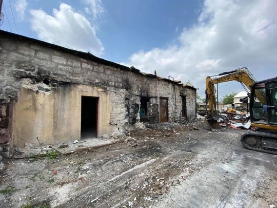 Damaged building, dark patches from fire, with excavator in the background, demolition taking place.