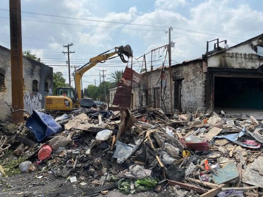 Demolition of charred brick building with excavator. Debris and rubble in foreground, blue sky above.