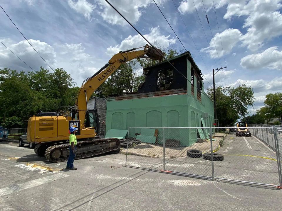 A yellow excavator demolishes a green building, smoke damage visible, under a cloudy sky.