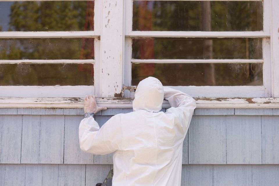 Abatement worker in a white protective suit inspecting a window frame on a wood-sided house