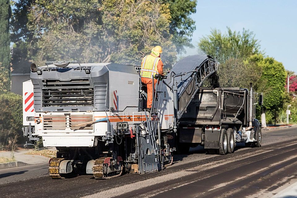 Worker on a cold planer milling asphalt off a road and loading it into a dump truck
