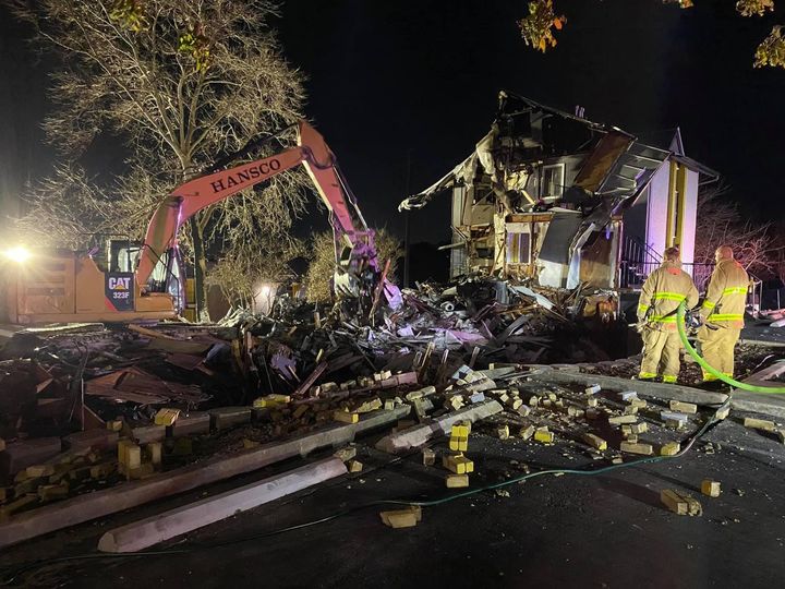 Firefighters hosing down debris as a Hansco excavator demolishes a burned house at night