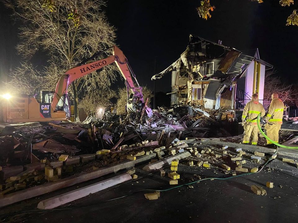 Firefighters hosing down debris as a Hansco excavator demolishes a burned house at night
