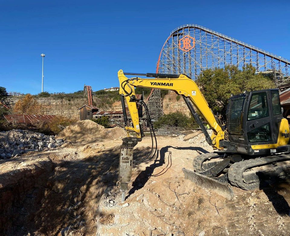 Yanmar mini excavator with a hydraulic breaker digging on a site beside a roller coaster