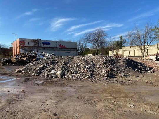 Pile of rubble in an empty lot, with a partially demolished brick building in the background under a blue sky.