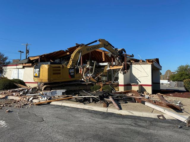 Excavator demolishing a single-story commercial building, with debris piled at the curb