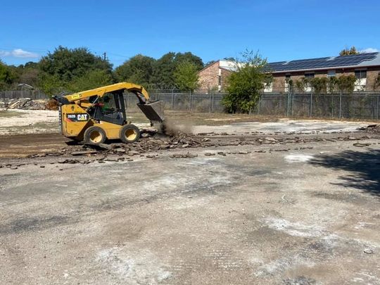 Yellow CAT skid steer clearing broken pavement from a fenced lot on a sunny day.