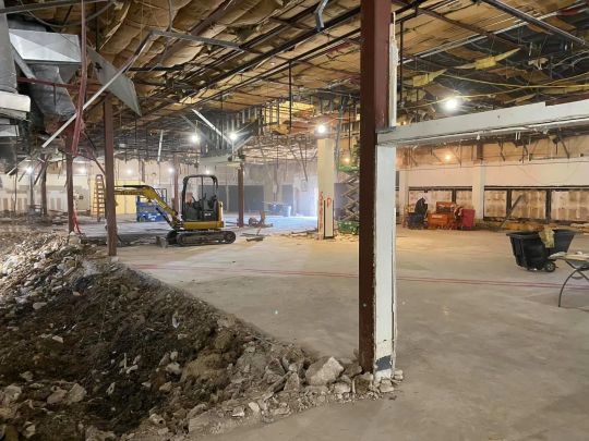 Construction site interior; excavator, debris, exposed structure; workers in the background.