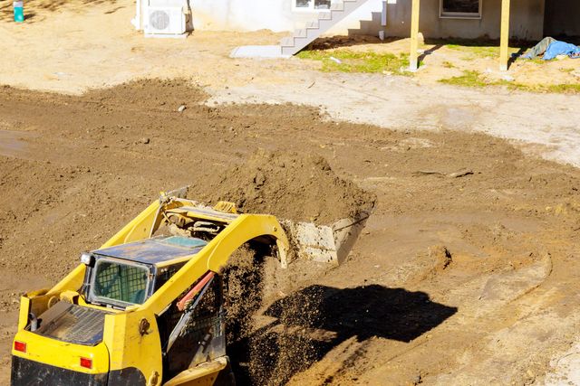 Yellow skid steer grading loose soil on a cleared lot after a building was demolished