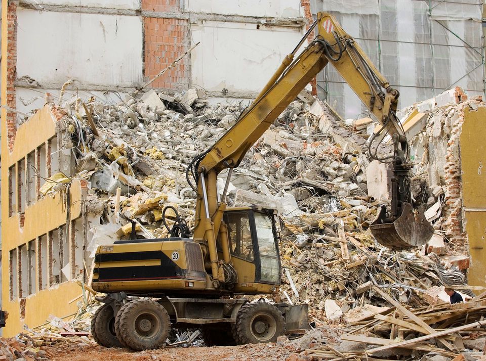 Wheeled excavator working a pile of rubble beside a partly demolished building