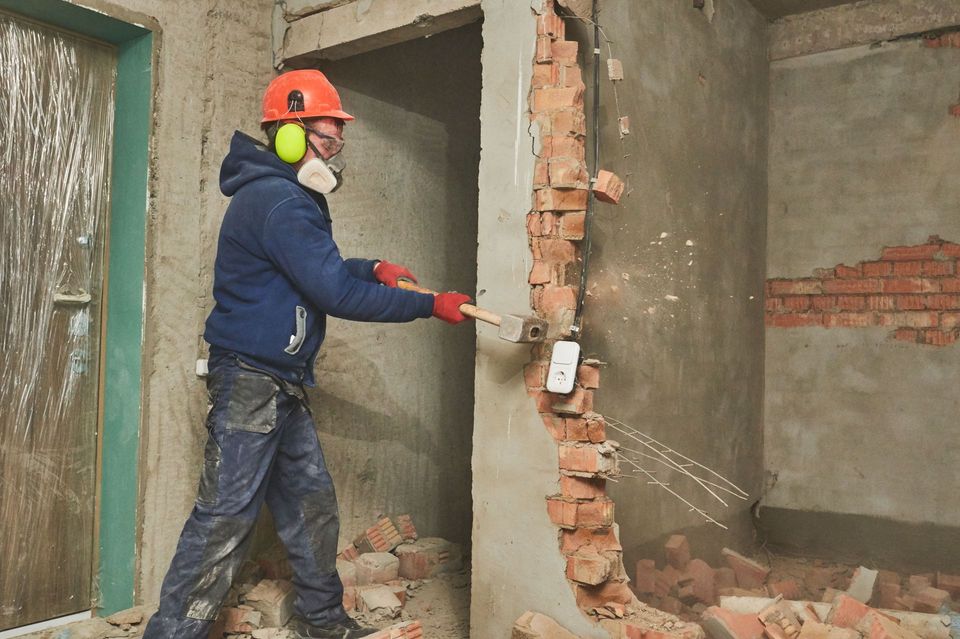 Worker in safety gear knocking out a brick interior wall with a sledgehammer