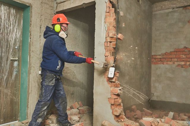 Worker in safety gear knocking out a brick interior wall with a sledgehammer